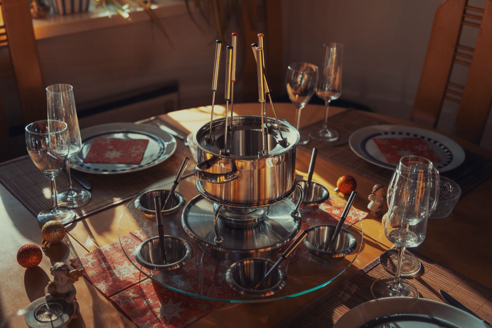 A stainless steel fondue set on a laid table with long forks, wine glasses and Christmas decorations
