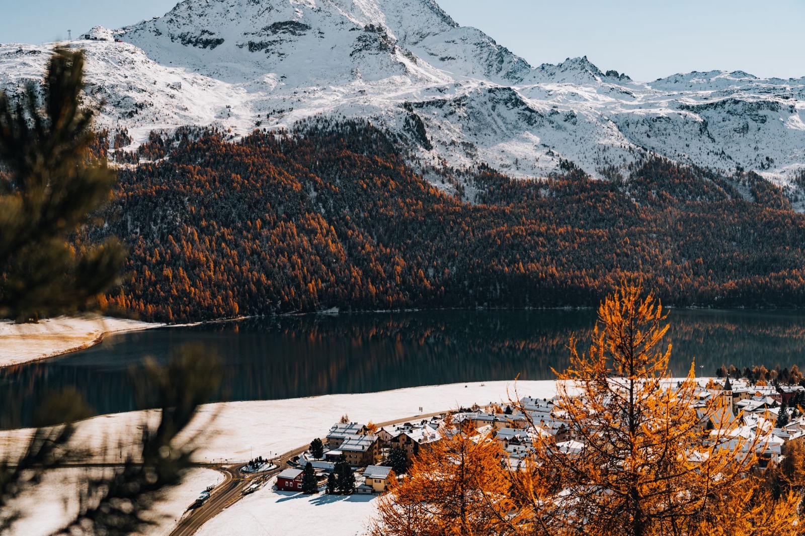 Golden larches and a snow-dusted village beside a dark lake below snowy peaks