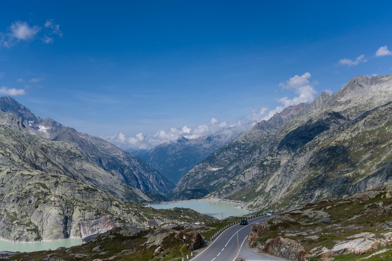 A two-lane mountain road descending past a pale turquoise reservoir between granite peaks