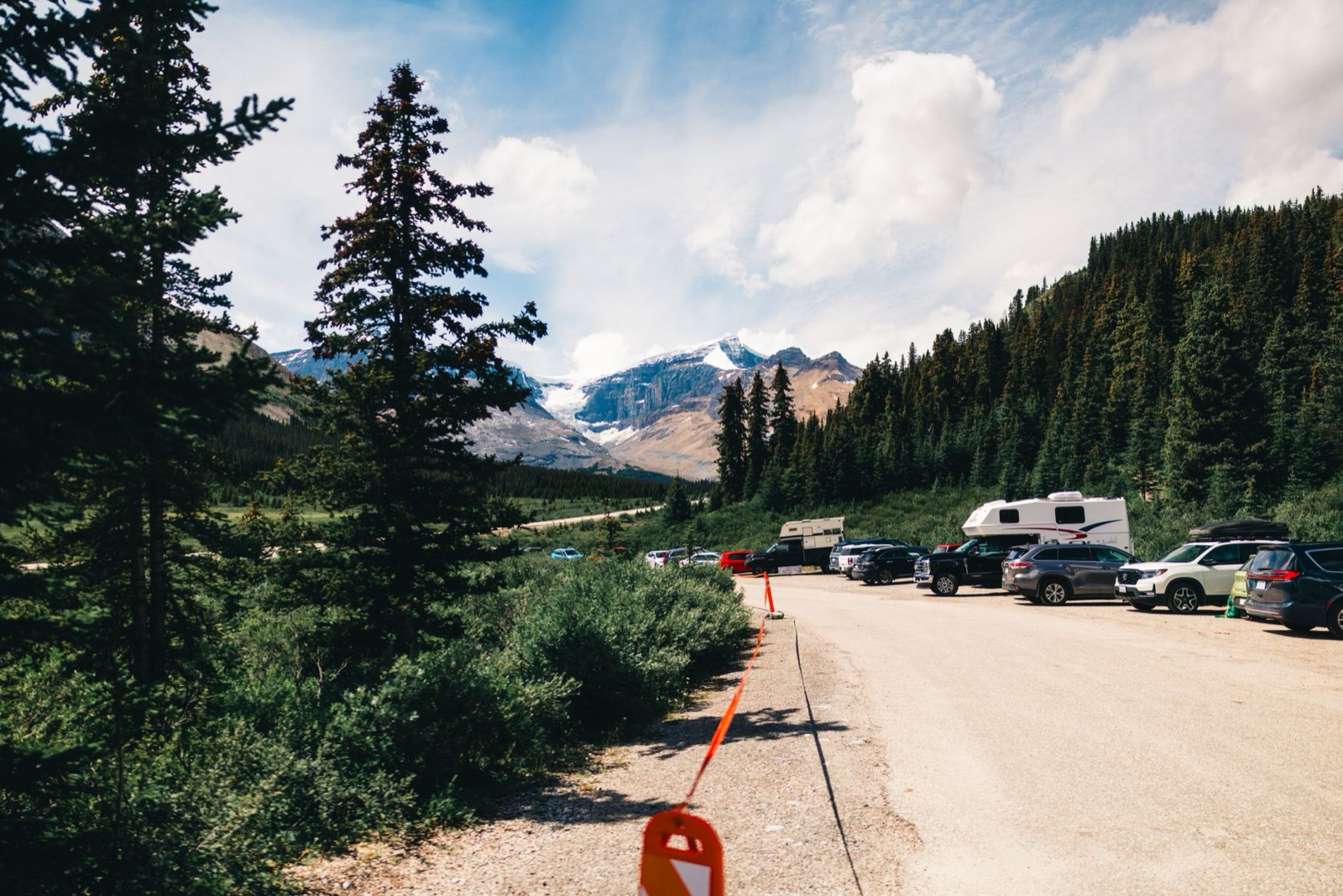 Cars and a white motorhome parked along a gravel car park below glaciated peaks