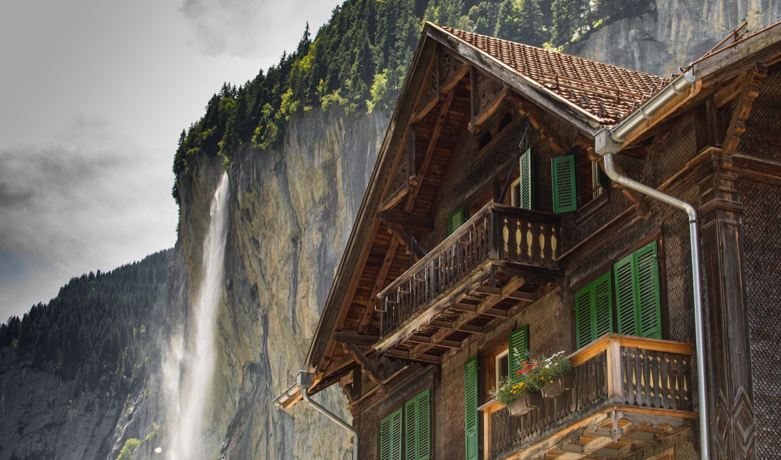 A wooden chalet with green shutters and flowered balconies below a cliff with a high waterfall